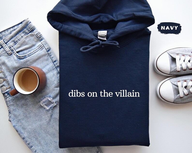 May include: A navy blue hoodie with the text "dibs on the villain" in white. The image also shows light blue jeans, a brown mug, and grey trainers.
