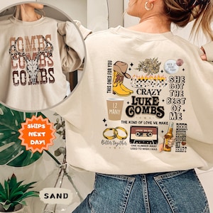 May include: Sand-colored sweatshirt featuring a graphic design with the text "COMBS" in a stacked layout, with a deer skull and antlers. The back of the sweatshirt has various graphics and text, including "LUKE COMBS" and "SHE GOT THE BEST OF ME."