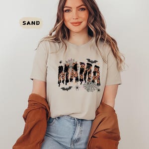 May include: A sand-colored t-shirt featuring the word "MAMA" in a leopard print design with black dripping accents. The shirt is decorated with spiderwebs, spiders, and bats, creating a Halloween theme.