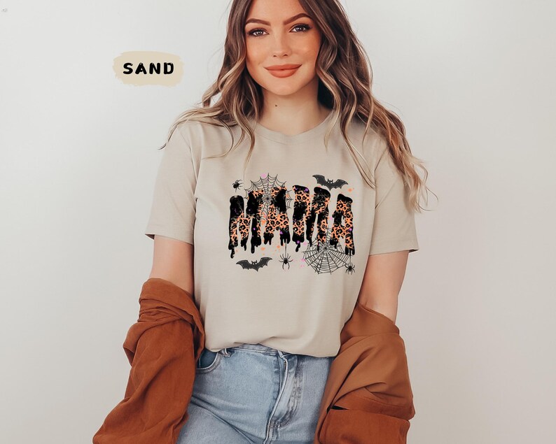 May include: A sand-colored t-shirt featuring the word "MAMA" in a leopard print design with black dripping accents. The shirt is decorated with spiderwebs, spiders, and bats, creating a Halloween theme.