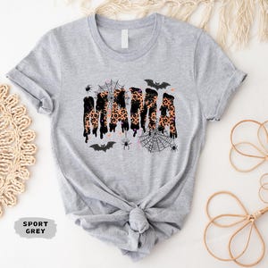 May include: Sport grey t-shirt with a Halloween-themed design. The word "MAMA" is printed in a dripping style with a leopard print pattern. The design includes bats, spiders, and spiderwebs.