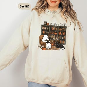 May include: Sand-colored hoodie featuring a Halloween-themed graphic. The graphic depicts a ghost reading in a cozy armchair in front of a bookshelf, with a black cat, pumpkins, and skulls. The word "SAND" is printed at the top.