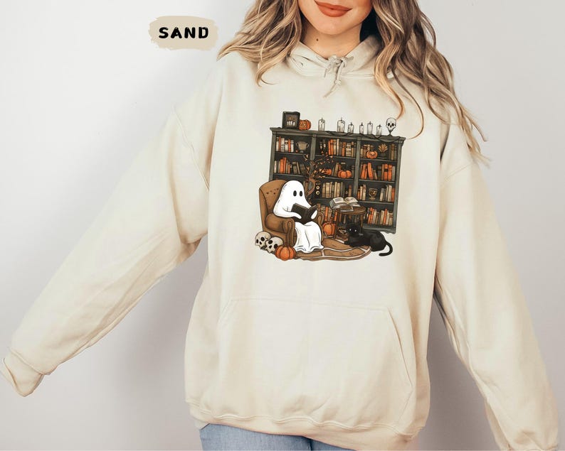May include: Sand-colored hoodie featuring a Halloween-themed graphic. The graphic depicts a ghost reading in a cozy armchair in front of a bookshelf, with a black cat, pumpkins, and skulls. The word "SAND" is printed at the top.