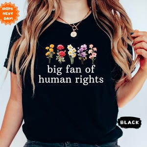 May include: Black t-shirt with the text "big fan of human rights" in white. The shirt features a floral design with yellow, red, white, purple, and pink flowers. The shirt is a crew neck and short-sleeved.