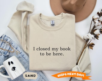 May include: Sand-colored sweatshirt with the text "I closed my book to be here." The sweatshirt is laid flat with a pair of blue jeans, a white ghost-shaped mug, and a brown hat. A sprig of cotton stems is also present.