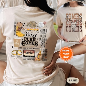 May include: Sand-colored t-shirt featuring a graphic design with the text "Luke Combs" and various country-themed illustrations. The design includes a cowboy hat, boots, a cassette tape, and beer bottle. The shirt also has the text "Better Together".