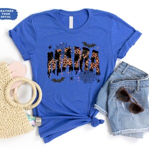 May include: A royal blue t-shirt with the word "MAMA" in a dripping leopard print design, accented with bats, spiders, and a web. The shirt is paired with denim shorts, sunglasses, and a straw bag.