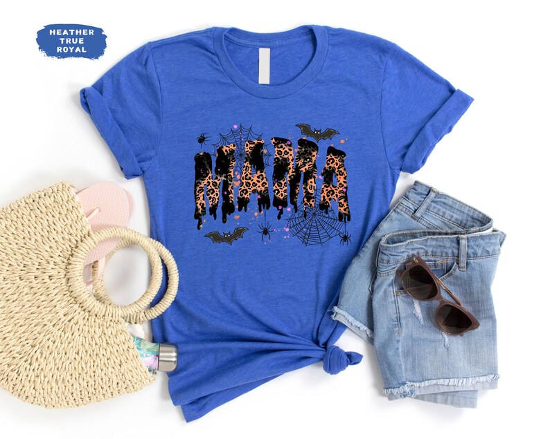 May include: A royal blue t-shirt with the word "MAMA" in a dripping leopard print design, accented with bats, spiders, and a web. The shirt is paired with denim shorts, sunglasses, and a straw bag.