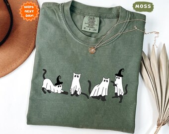 May include: Moss green t-shirt featuring a playful design of four cartoon cats dressed as ghosts, each wearing a witch hat. The shirt has a crew neck and short sleeves. A delicate gold necklace with a pendant rests on the shirt. The tag reads "Comfort Colors".
