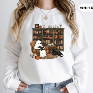 May include: White sweatshirt with a Halloween-themed graphic. The graphic depicts a ghost reading a book in a cozy armchair in front of a bookshelf. A black cat, pumpkins, and skulls complete the scene. The word "WHITE" is printed on the top right.