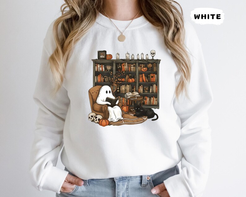 May include: White sweatshirt with a Halloween-themed graphic. The graphic depicts a ghost reading a book in a cozy armchair in front of a bookshelf. A black cat, pumpkins, and skulls complete the scene. The word "WHITE" is printed on the top right.