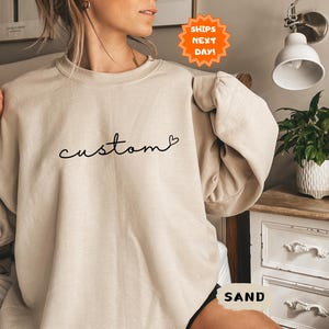May include: A beige sweatshirt with the word "custom" written in black script with a small heart symbol. The sweatshirt has a relaxed fit and a crew neck.