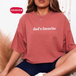 May include: A coral-colored t-shirt with the text "dad's favorite" in white. The shirt has short sleeves and a relaxed fit. The Crimson logo is visible in the upper left corner.