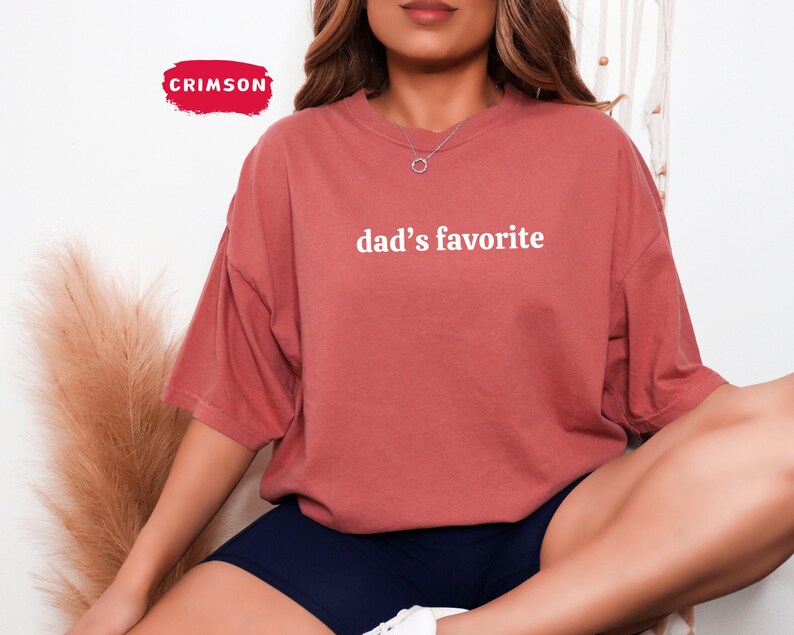 May include: A coral-colored t-shirt with the text "dad's favorite" in white. The shirt has short sleeves and a relaxed fit. The Crimson logo is visible in the upper left corner.