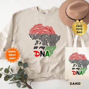 May include: Sand-colored sweatshirt with a red, black, and green fingerprint design in the shape of Africa, with the text "It's in my DNA." A matching tote bag and a brown hat are also visible.