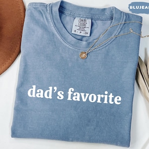 May include: A blue jean-colored t-shirt with the words "dad's favorite" printed in white. The shirt has a crew neck and short sleeves. A gold necklace with a pendant is draped across the shirt.