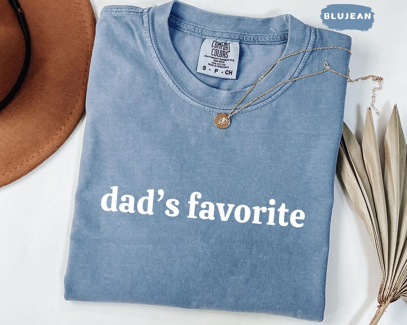 May include: A blue jean-colored t-shirt with the words "dad's favorite" printed in white. The shirt has a crew neck and short sleeves. A gold necklace with a pendant is draped across the shirt.