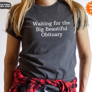 May include: Heather dark grey t-shirt with the text "Waiting for the Big Beautiful Obituary" in white. The shirt is a crew neck style. A red and black plaid shirt is tied around the waist.