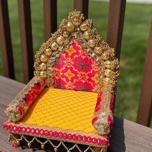 May include: A decorative, ornate miniature chair with a yellow seat and back, and red and gold patterned fabric. The chair is trimmed with gold beads and sequins, and has two rolled armrests.