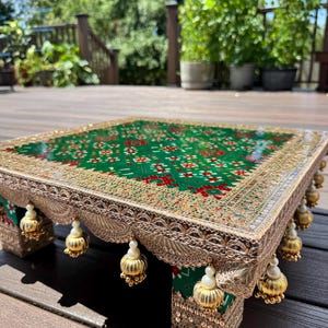 Può includere: Un tavolino quadrato a profilo basso caratterizzato da una superficie verde brillante con un motivo geometrico rosso e bianco. I bordi sono decorati con intricate finiture metalliche dorate e ornamenti sferici dorati pendenti con perline bianche.