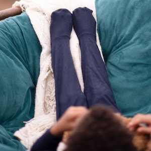 May include: A person wearing dark blue tights is relaxing in a teal beanbag chair with a white blanket. The person is holding a stuffed animal.