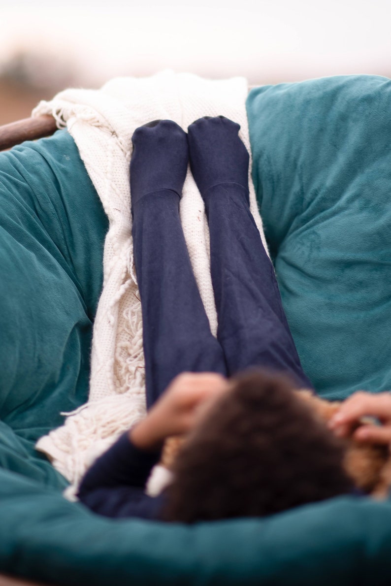 May include: A person wearing dark blue tights is relaxing in a teal beanbag chair with a white blanket. The person is holding a stuffed animal.