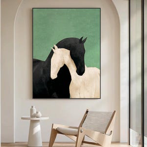 May include: A framed art print featuring two horses. One horse is black, and the other is white. The background is a solid green color. The artwork is displayed in a modern interior setting with a white side table and a wooden chair.