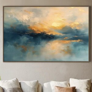 May include: Large framed abstract painting with a sunset over water theme. The artwork features shades of blue, gold, and white, creating a serene and calming atmosphere. The painting is displayed above a cream-colored sectional sofa.