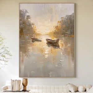 May include: An oil painting depicts a serene lake scene with two small boats. The artwork features a muted color palette of grays, browns, and yellows, with reflections in the water. The painting is framed and hangs above a white sofa.