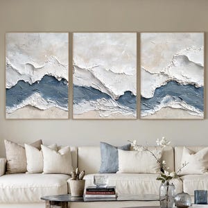 May include: A triptych of abstract wall art featuring textured, layered designs. Each piece has a neutral beige and white background with a deep blue accent, creating a coastal or landscape theme. The artwork is framed in a light wood.