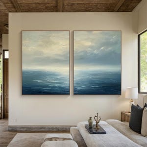 May include: Two framed abstract ocean paintings. The artwork features a seascape with shades of blue and white, depicting the sky and water. The paintings are displayed on a neutral-colored wall in a well-lit room, with a sofa and decor in the foreground.