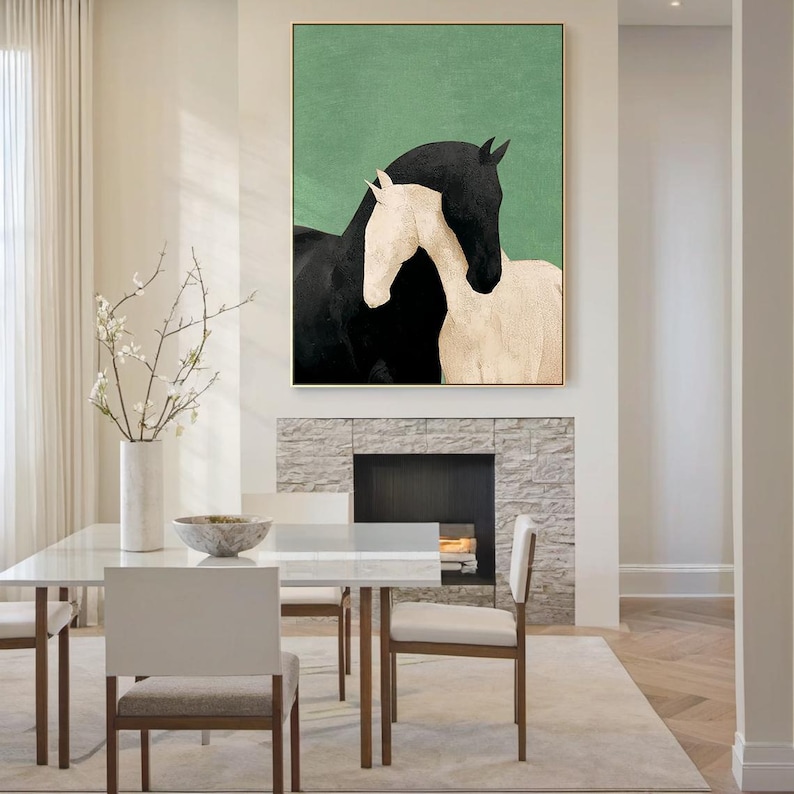 May include: Abstract art print featuring two horses, one black and one cream-colored, against a green backdrop. The artwork is framed in gold and hangs above a fireplace. The room has a modern, minimalist design with a white dining table and chairs.