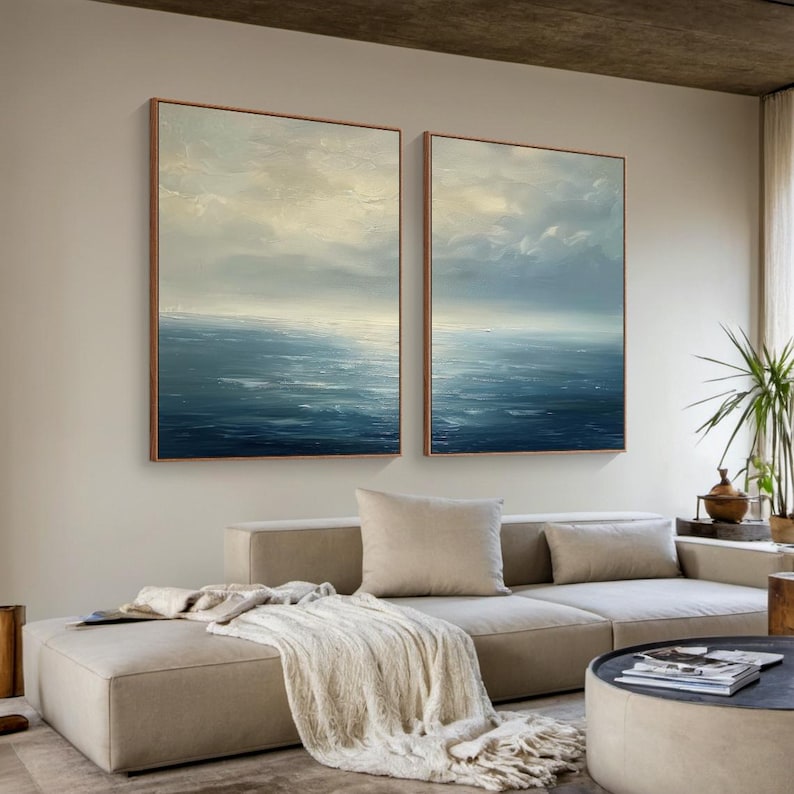 May include: Two framed abstract seascape paintings. The artwork features a textured, layered effect with shades of blue, white, and beige, evoking a sense of the ocean and sky. The frames are a warm brown color. The paintings are displayed above a beige sectional sofa.