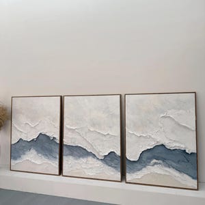 May include: A triptych of three rectangular wall art pieces featuring heavy white and blue impasto texture. Each canvas displays a layered, abstract landscape design with thick, raised white paint strokes contrasting against a deep blue base.