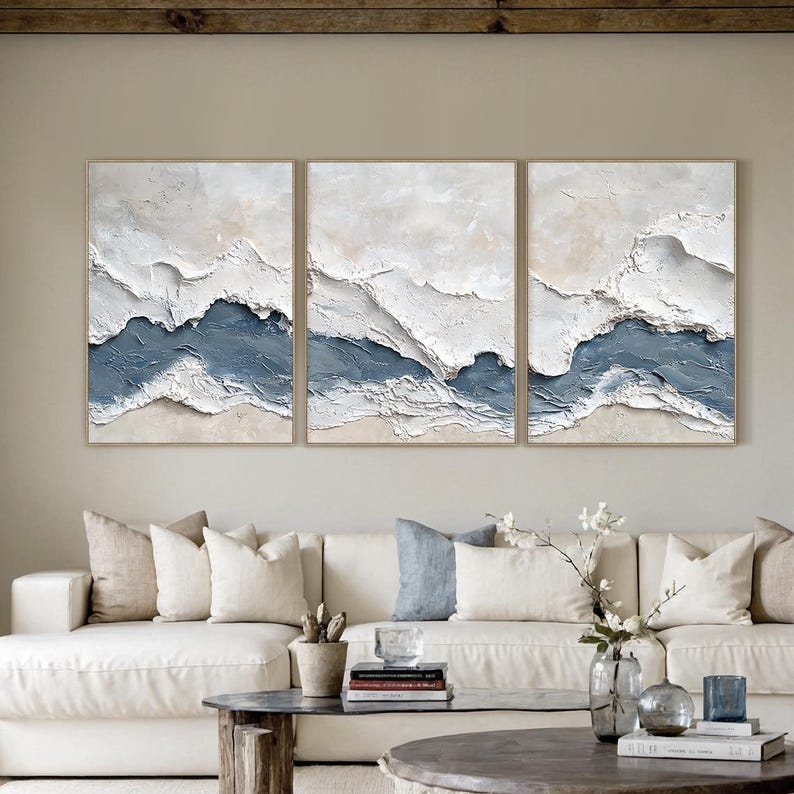 May include: A triptych of abstract wall art featuring textured, layered designs. Each piece has a neutral beige and white background with a deep blue accent, creating a coastal or landscape theme. The artwork is framed in a light wood.