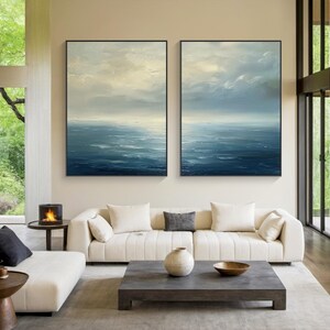 May include: Two framed abstract ocean paintings. The artwork features a textured, layered effect with shades of blue, white, and gray, depicting a seascape with a cloudy sky. The paintings are displayed above a white sofa in a modern living room.