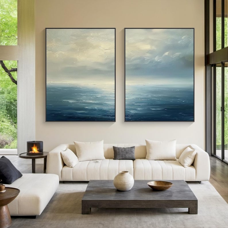 May include: Two framed abstract ocean paintings. The artwork features a textured, layered effect with shades of blue, white, and gray, depicting a seascape with a cloudy sky. The paintings are displayed above a white sofa in a modern living room.