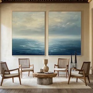 May include: Two framed abstract paintings depict a seascape with shades of blue and white, evoking a sense of calm. The paintings hang above a living room setting with wooden chairs, a coffee table, and decorative items.