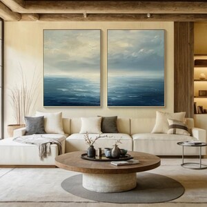 May include: Two framed abstract ocean paintings with gold frames hang above a cream-colored sectional sofa. The paintings depict a seascape with blue and white hues. A round wooden coffee table with decorative items sits in front of the sofa, on a gray rug.