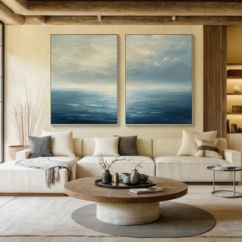 May include: Two framed abstract ocean paintings with gold frames hang above a cream-colored sectional sofa. The paintings depict a seascape with blue and white hues. A round wooden coffee table with decorative items sits in front of the sofa, on a gray rug.