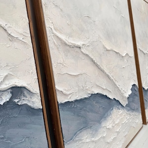 May include: A triptych of three rectangular wall art panels framed in dark wood. Each panel features a thick, textured impasto application of white and slate blue paint, creating raised, layered ridges that mimic a landscape or abstract terrain.