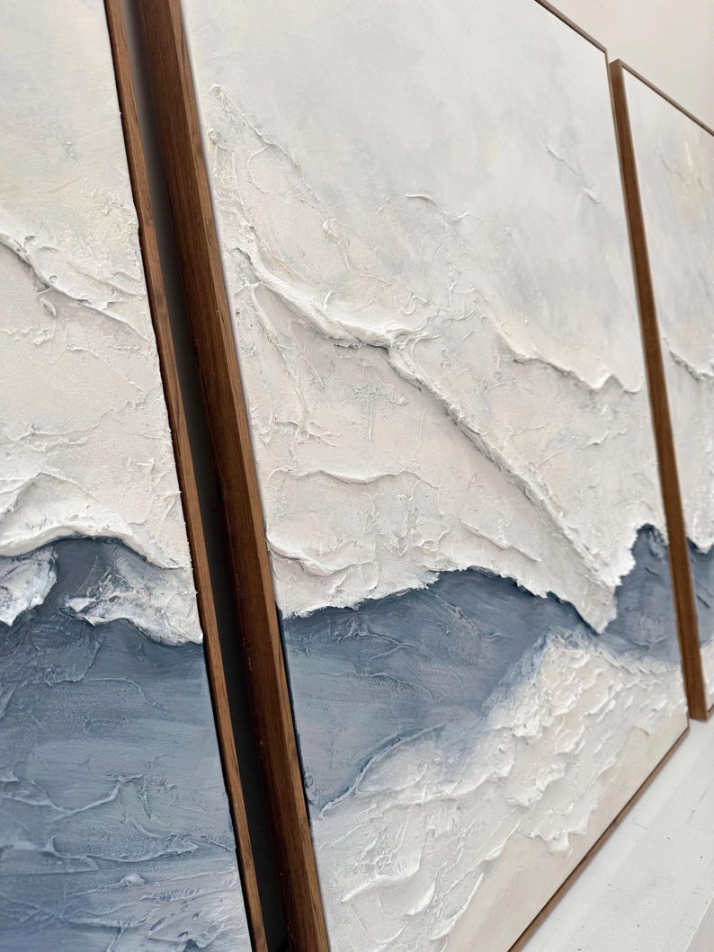 May include: A triptych of three rectangular wall art panels framed in dark wood. Each panel features a thick, textured impasto application of white and slate blue paint, creating raised, layered ridges that mimic a landscape or abstract terrain.
