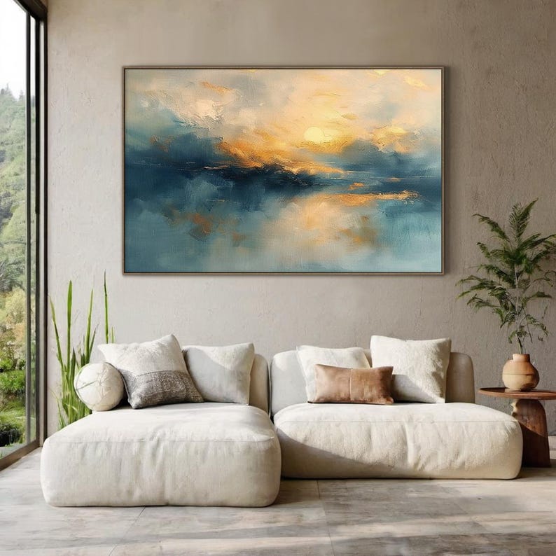 May include: Large framed abstract painting with a sunset over water theme. The artwork features shades of blue, gold, and white, creating a serene and calming atmosphere. The painting is displayed above a cream-colored sectional sofa.
