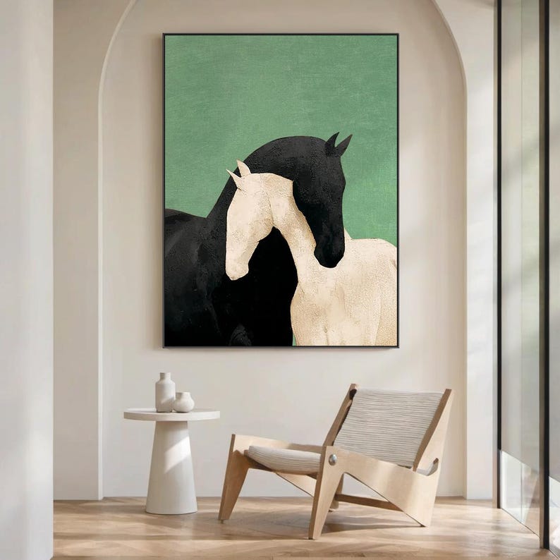May include: A framed art print featuring two horses. One horse is black, and the other is white. The background is a solid green color. The artwork is displayed in a modern interior setting with a white side table and a wooden chair.