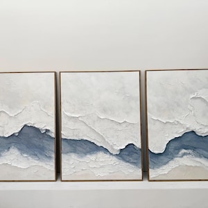 May include: A triptych of three rectangular wall art pieces featuring heavy white and blue impasto texture. Each canvas displays layered, raised white paint strokes over a deep blue base, creating a landscape effect within thin wooden frames.