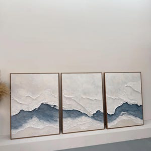 May include: A triptych of three rectangular wall art pieces featuring thick, textured white impasto paint layered over deep blue and beige tones. Each canvas is framed in a thin, light-toned wooden border, creating a cohesive landscape-inspired composition with raised, sculptural ridges across the surface.