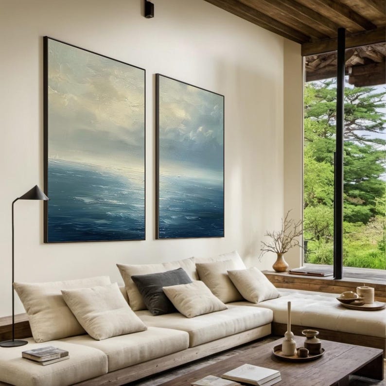 May include: Two framed abstract seascape paintings in shades of blue and white hang on a cream-colored wall. The paintings are above a beige sectional sofa with several pillows. A modern black floor lamp is to the left of the paintings.