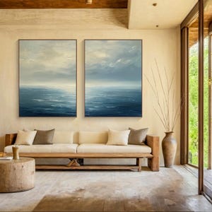 May include: Two framed abstract ocean paintings in shades of blue and white hang above a wooden sofa with neutral-colored cushions. The room has a natural, earthy aesthetic with a wooden coffee table and a large vase.
