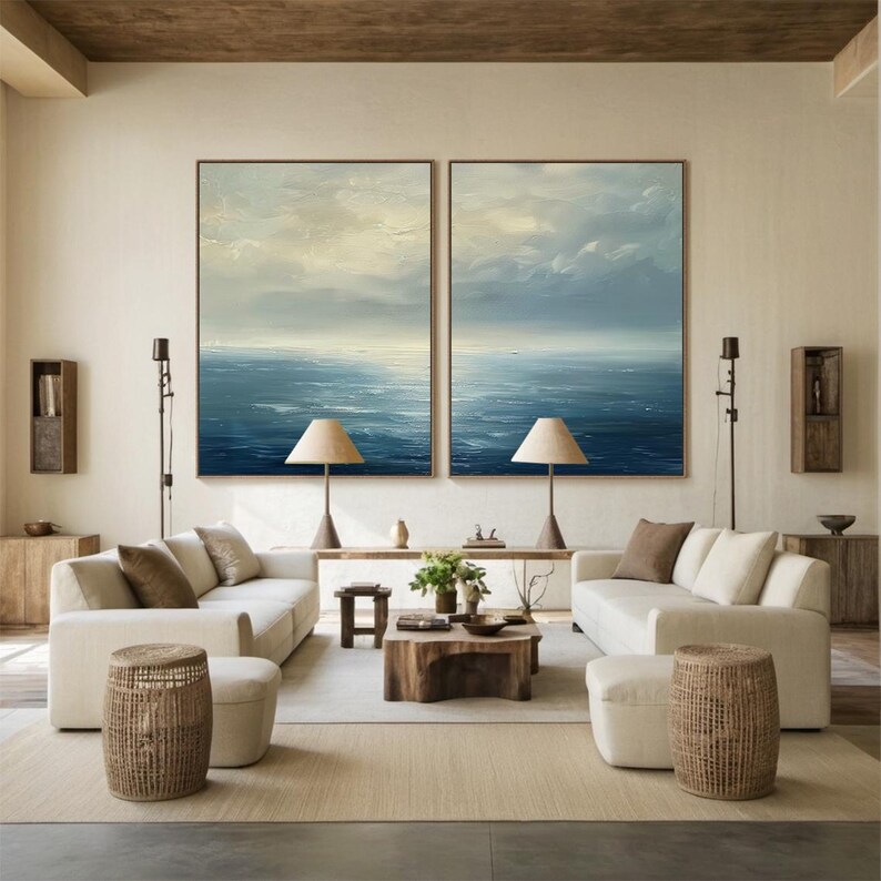May include: A living room with two large framed paintings of the ocean. The room features two white sofas, wooden tables, and woven stools. Two lamps with beige shades sit on the tables. The color palette is neutral with beige, brown, and blue tones.