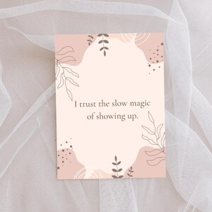 Könnte beinhalten: Eine rechteckige Karte mit einem zartrosa Hintergrund und abstrakten Formen in Weiß und Braun. Die Karte enthält den Text "I trust the slow magic of showing up." Dekorative Blattillustrationen umrahmen den Text.